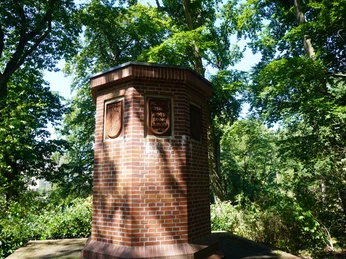 Ein aus rotem Backstein errichtetes Mahnmal mit Wappen und Inschrift, umgeben von Bäumen im Grünen.