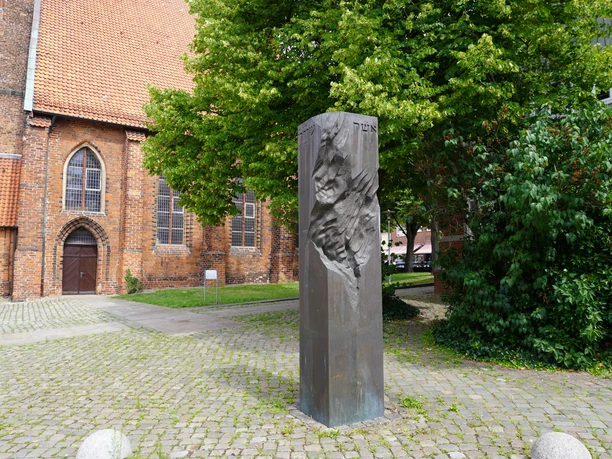 Mahnsäule aus dunklem Stein mit einer Reliefstruktur, umgeben von Bäumen und Backsteingebäude.