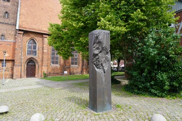 Mahnmal für die jüdischen Opfer Verden Mahnsäule aus dunklem Stein mit einer Reliefstruktur, umgeben von Bäumen und Backsteingebäude.