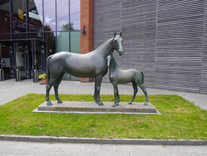 Bronzeskulptur einer Pferdestute mit ihrem stehenden Fohlen auf einem Sockel vor einem modernen Gebäude.