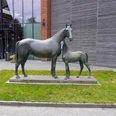 Skulptur Mutterstute mit Fohlen Verden Bronzeskulptur einer Pferdestute mit ihrem stehenden Fohlen auf einem Sockel vor einem modernen Gebäude.
