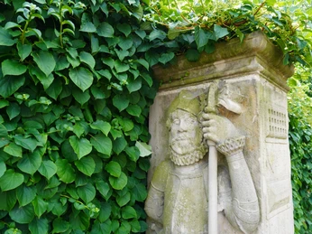 Wächterfiguren Verden Steinerne Wächterfigur aus Verden, halb in dichte grüne Blätter eingehüllt, mit reich verziertem Bart.