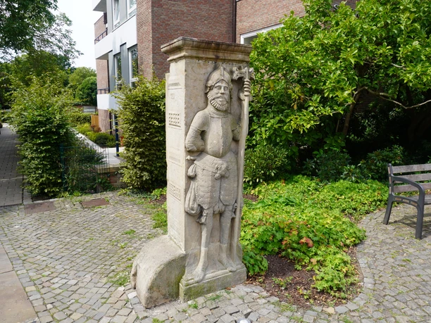 Steinskulptur eines mittelalterlichen Wächters in Verden, umgeben von Grün und Pflastersteinen.