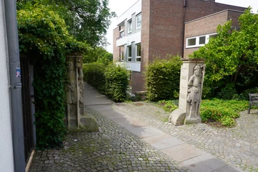 Wächterfiguren Verden Zwei steinerne Wächterfiguren flankieren ein Kopfsteinpflaster auf dem Weg zu einem modernen Backsteingebäude.