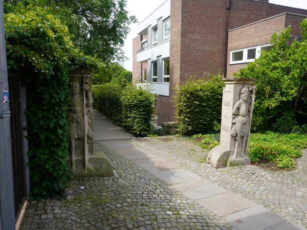 Wächterfiguren Verden Zwei steinerne Wächterfiguren flankieren ein Kopfsteinpflaster auf dem Weg zu einem modernen Backsteingebäude.