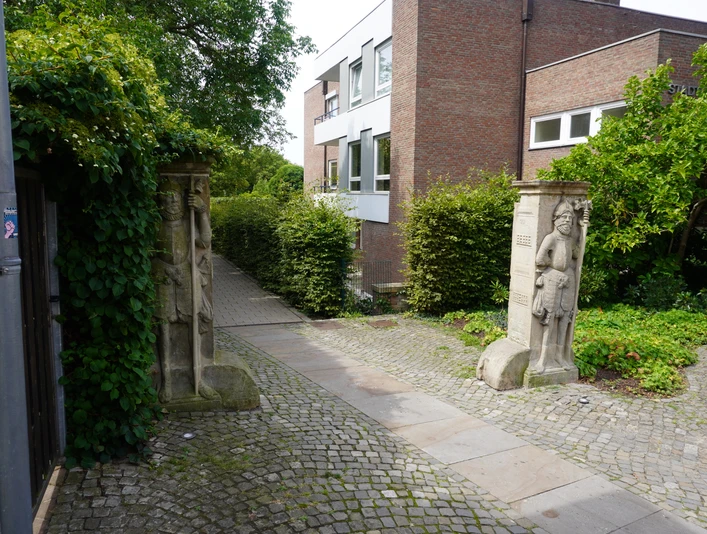 Wächterfiguren Verden Zwei steinerne Wächterfiguren flankieren ein Kopfsteinpflaster auf dem Weg zu einem modernen Backsteingebäude.
