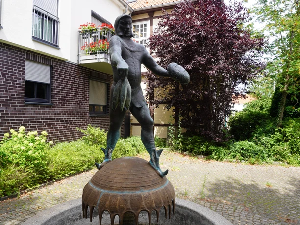 Brunnen mit einer Statue des Seeräubers Störtebeker in einer ruhigen Gartenanlage an einem gepflasterten Weg.