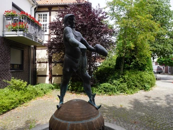 Bronzestatue eines Piraten mit Schwert, umgeben von Hecken und Gebäuden in einer gepflasterten Passagen.