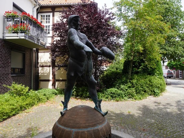 Bronzestatue eines Piraten mit Schwert, umgeben von Hecken und Gebäuden in einer gepflasterten Passagen.
