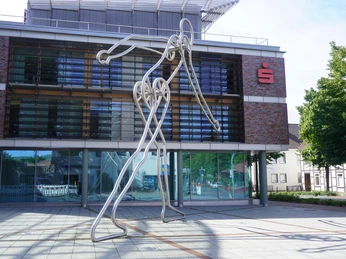 Die Skulptur "Walking Man" aus Metallstäben steht vor einer modernen Bank mit großen Glasfenstern.