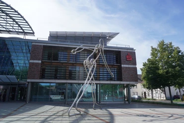 Walking Man Verden Die Skulptur Walking Man aus Metall steht vor einem modernen Gebäude mit Glasfassade in Verden.
