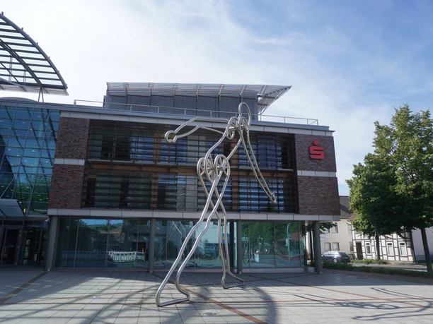 Walking Man Verden Die Skulptur Walking Man aus Metall steht vor einem modernen Gebäude mit Glasfassade in Verden.