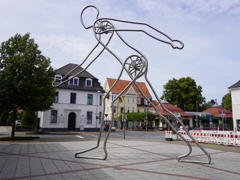 Moderne Metallskulptur eines gehenden Mannes in Verden, umgeben von Gebäuden und Bäumen.