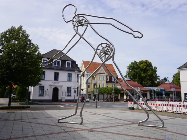 Skulptur Walking Man Verden Moderne Metallskulptur eines gehenden Mannes in Verden, umgeben von Gebäuden und Bäumen.