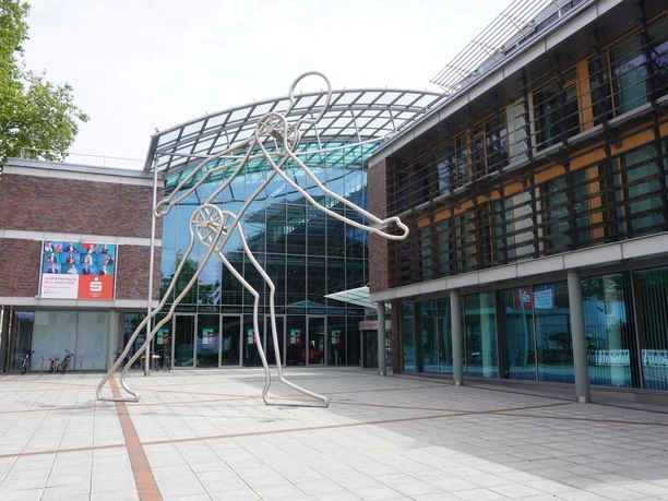 Eine große, moderne Skulptur eines gehenden Mannes aus Metall steht vor einem gläsernen Gebäude.