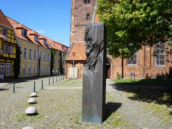 Denkmal aus Bronze nahe einer Kirche, umgeben von historischen Gebäuden, in hellem Sonnenlicht.