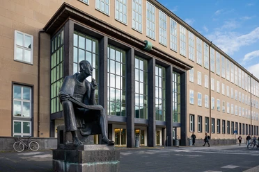 Albertus Magnus Denkmal Das Albertus-Magnus-Denkmal, eine bronzene Statue des sitzenden Gelehrten, vor einer modernen Gebäudeansicht in Köln.