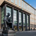 Albertus Magnus Denkmal Das Albertus-Magnus-Denkmal, eine bronzene Statue des sitzenden Gelehrten, vor einer modernen Gebäudeansicht in Köln.The Albertus Magnus monument, a bronze statue of the seated scholar, in front of a modern building in Cologne.