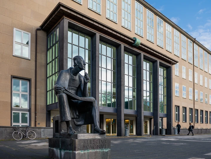 Albertus Magnus Denkmal Das Albertus-Magnus-Denkmal, eine bronzene Statue des sitzenden Gelehrten, vor einer modernen Gebäudeansicht in Köln.The Albertus Magnus monument, a bronze statue of the seated scholar, in front of a modern building in Cologne.