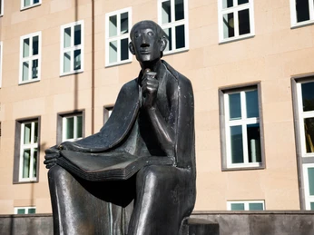 Albertus Magnus Denkmal Das Albertus-Magnus-Denkmal zeigt eine sitzende Bronzestatue vor einem Gebäude, ein Buch haltend, grübelnd.The Albertus Magnus monument shows a seated bronze statue in front of a building, holding a book and pondering.