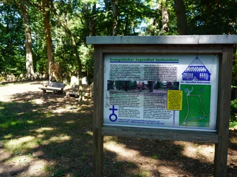 Holzrahmen-Infotafel im Sachsenhain Verden, umgeben von schattenspendenden Bäumen und Sitzgelegenheit.