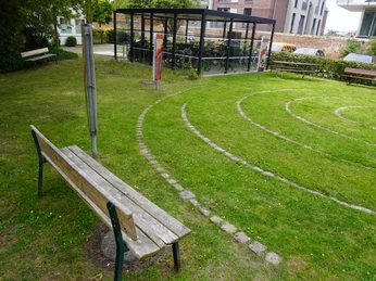 Park mit gepflegtem Spirallabyrinth aus Steinen, umgeben von grüner Rasenfläche und Bänken.