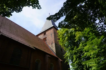St. Andreaskirche Backsteinkirche mit hohem Turm und spitzem Dach, umgeben von grünen Bäumen, vor blauem Himmel.
