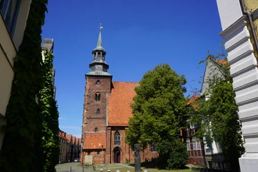 Backsteinkirche mit historischem Turm inmitten alter Gebäude, umgeben von Bäumen unter strahlend blauem Himmel.