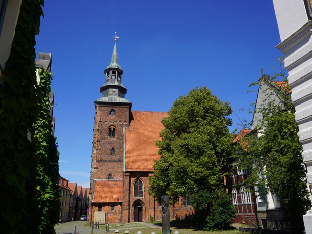 Backsteinkirche mit historischem Turm inmitten alter Gebäude, umgeben von Bäumen unter strahlend blauem Himmel.