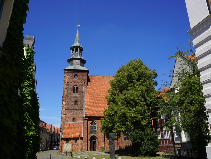 Backsteinkirche mit historischem Turm inmitten alter Gebäude, umgeben von Bäumen unter strahlend blauem Himmel.