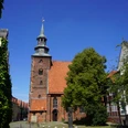 St. Johanniskirche Backsteinkirche mit historischem Turm inmitten alter Gebäude, umgeben von Bäumen unter strahlend blauem Himmel.