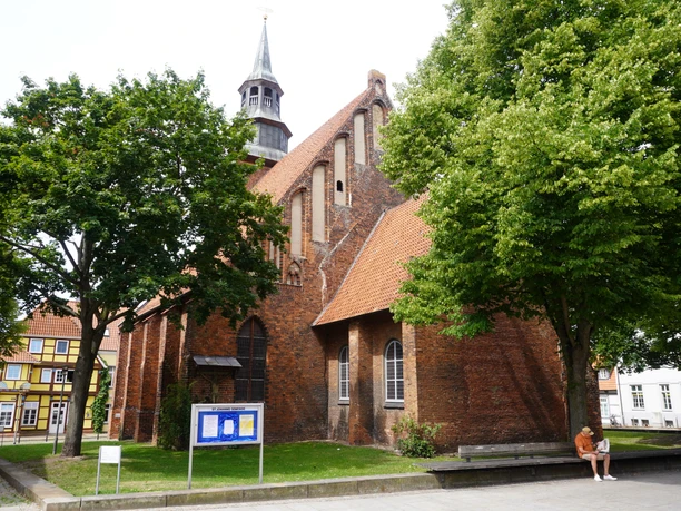 St. Johanniskirche Verden Backsteinkirche mit markantem Glockenturm, umgeben von Bäumen. Eine Person sitzt im Vordergrund.