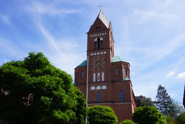 Backsteinkirche mit grünem Dach, Glockenturm und strahlend blauem Himmel, umgeben von Bäumen.
