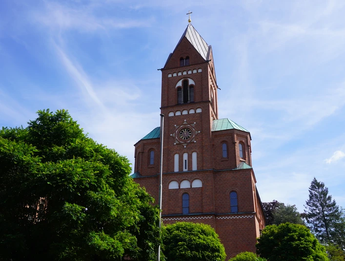 Backsteinkirche mit grünem Dach, Glockenturm und strahlend blauem Himmel, umgeben von Bäumen.