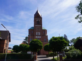 Historische Backsteinkirche mit hohem Turm und Kreuz, umgeben von Bäumen und blauem Himmel.