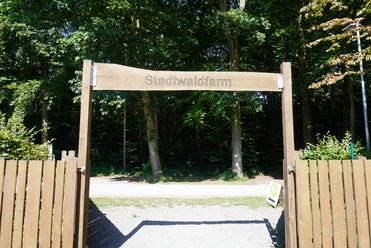 Eingang zur Stadtwaldfarm Verden mit hölzernem Torbogen, umgeben von grünen Bäumen im Hintergrund.