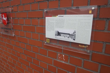 Amtsgericht Eine Infotafel zum Stiftshof Verden an einer roten Ziegelwand mit Niedersachsen-Wappen.