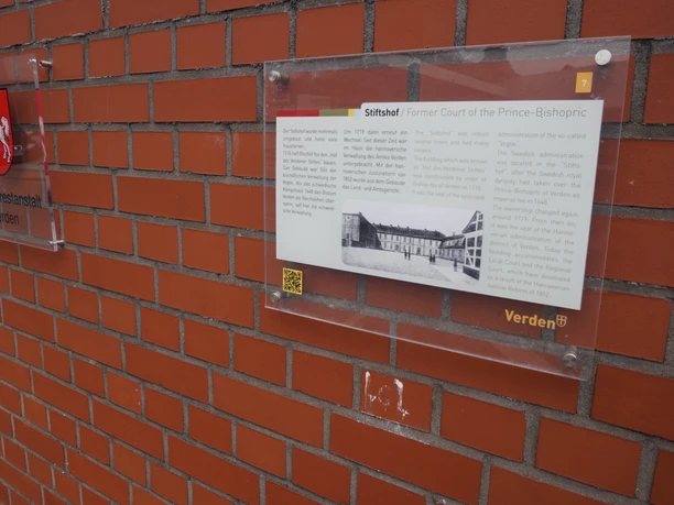 Amtsgericht Eine Infotafel zum Stiftshof Verden an einer roten Ziegelwand mit Niedersachsen-Wappen.
