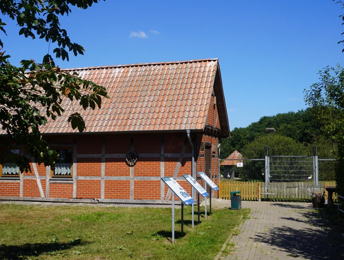 Storchenpflegestation in Verden mit Fachwerkgebäude, Infotafeln und grünem Umfeld unter blauem Himmel.