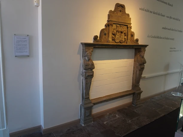 Stone relief featuring a coat of arms and figures at the Domherrenhaus Museum, displayed on a wall alongside a text passage.