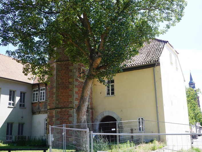 Ein großes Gebäude mit Backsteinmauer und Torbogen, flankiert von einem hohen Baum und Zaun.