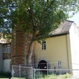 Syndikatshof Ein großes Gebäude mit Backsteinmauer und Torbogen, flankiert von einem hohen Baum und Zaun.