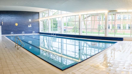 Schwimmbad Hesel Du erblickst das helle Hallenbad Hesel, dessen Wasser die breite Fensterfront klar reflektiert.