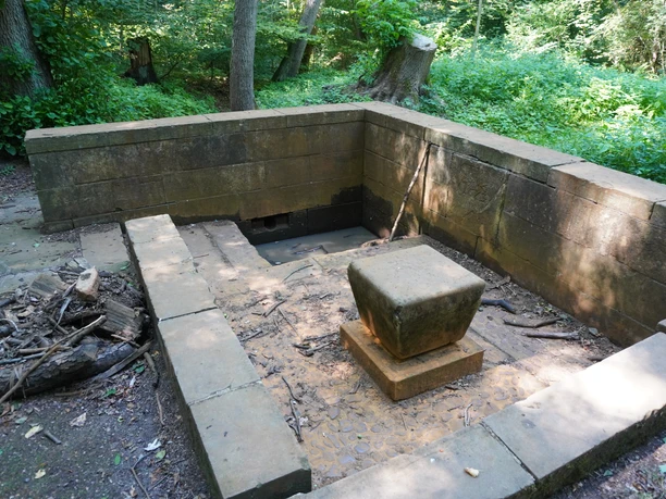 Brunnen Verden Ein steinerner Brunnen im Wald mit quadratischem Sockel, umgeben von Bäumen und dichtem Grün.