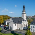 Saint Clemens Das Bild zeigt die Sankt Clemens Kirche in einer luftigen Perspektive, eingebettet in eine grüne städtische Umgebung. Die weiße Kirche mit schwarzem Dach und charakteristischem Turm steht neben einem historischen Gebäude bei sonnigem Wetter.The picture shows St. Clement's Church from an airy perspective, embedded in a green urban environment. The white church with its black roof and characteristic tower stands next to a historic building in sunny weather.