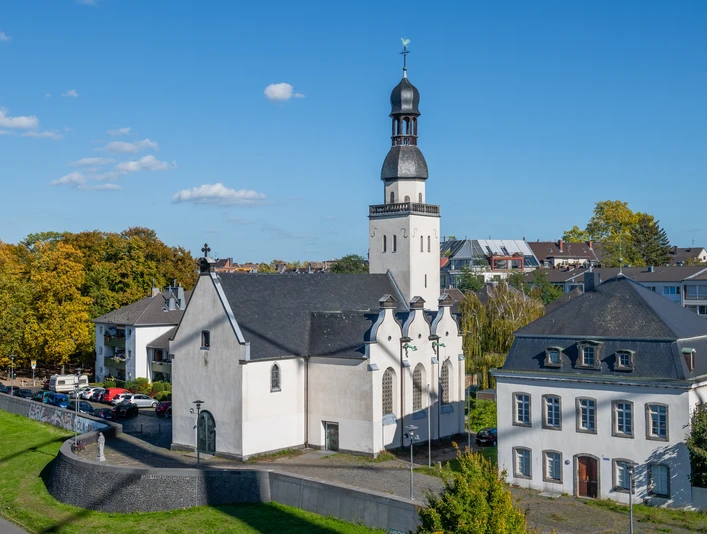 Saint Clemens Das Bild zeigt die Sankt Clemens Kirche in einer luftigen Perspektive, eingebettet in eine grüne städtische Umgebung. Die weiße Kirche mit schwarzem Dach und charakteristischem Turm steht neben einem historischen Gebäude bei sonnigem Wetter.The picture shows St. Clement's Church from an airy perspective, embedded in a green urban environment. The white church with its black roof and characteristic tower stands next to a historic building in sunny weather.