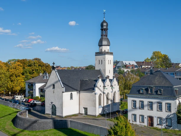 Sankt Clemens Das Bild zeigt die Sankt Clemens Kirche in einer luftigen Perspektive, eingebettet in eine grüne städtische Umgebung. Die weiße Kirche mit schwarzem Dach und charakteristischem Turm steht neben einem historischen Gebäude bei sonnigem Wetter.