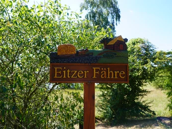 Holzwegweiser mit der Aufschrift "Eitzer Fähre", dekoriert mit Landschaft und Mühle, umgeben von Bäumen.
