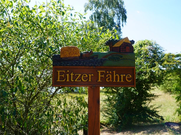 Eitzer Wegweiser Holzwegweiser mit der Aufschrift "Eitzer Fähre", dekoriert mit Landschaft und Mühle, umgeben von Bäumen.