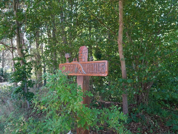 Holzwegweiser im Grünen markiert den Eitzer Mühle Pfad. Umgeben von Bäumen und dichten Büschen.